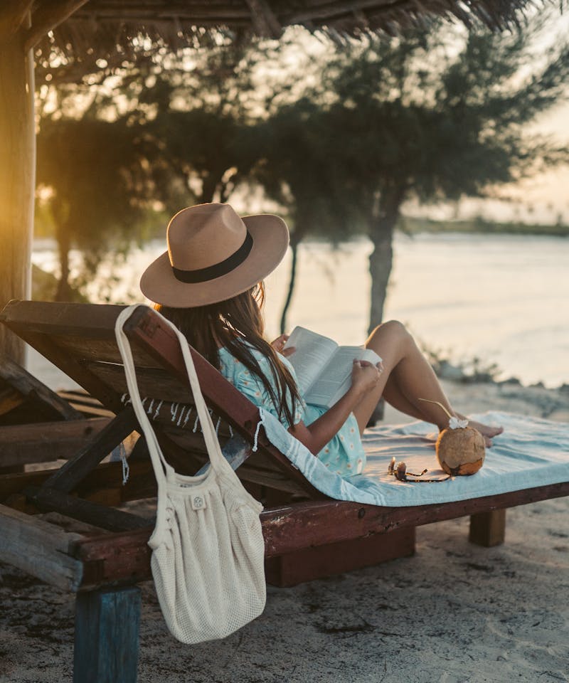 Woman reading on a beach lounger under a sun hat in Zanzibar at sunset, embracing vacation tranquility.